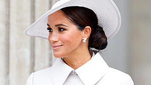 A close-up shot of Meghan Markle, wearing a wide wide-brimmed hat with her hair in a low chignon, with diamond drop earrings and a white collared trench