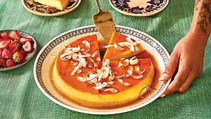 A coconut flan with shavings of coconut on top drizzled with caramel sauce on a white plate with a brown patterned border with a slice cut out of it and a vintage cake server lifting a slice up on a teal tablecoth