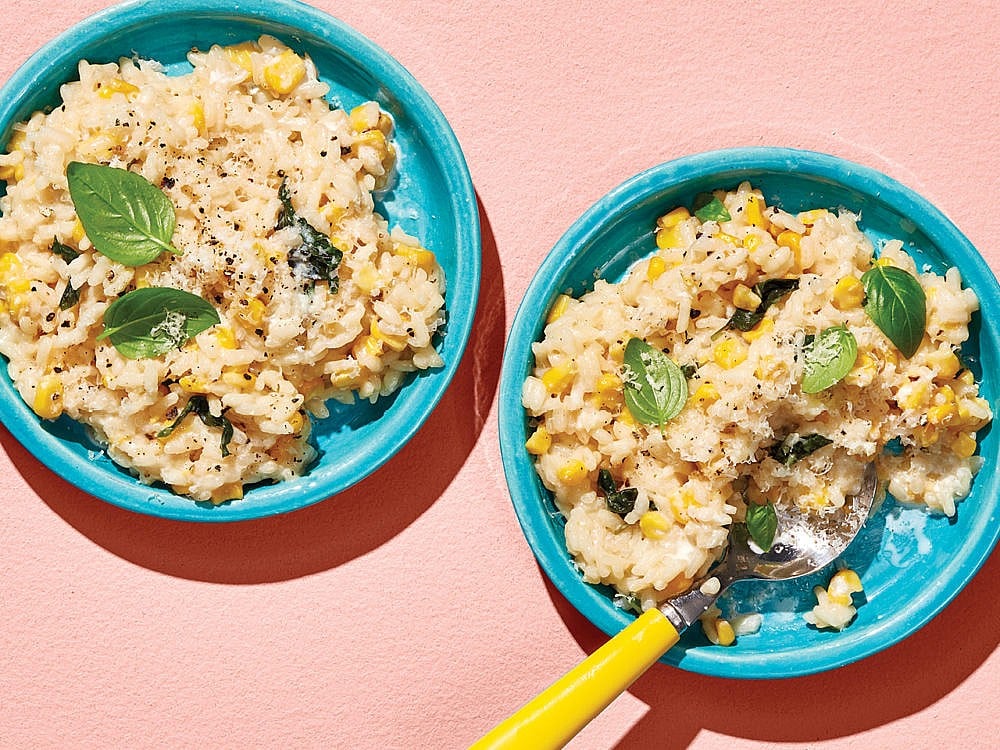 Two bowls of corn risotto topped with basil leaves against a pale pink background
