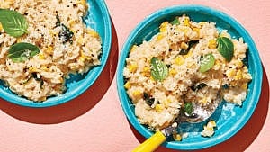 Two bowls of corn risotto topped with basil leaves against a pale pink background