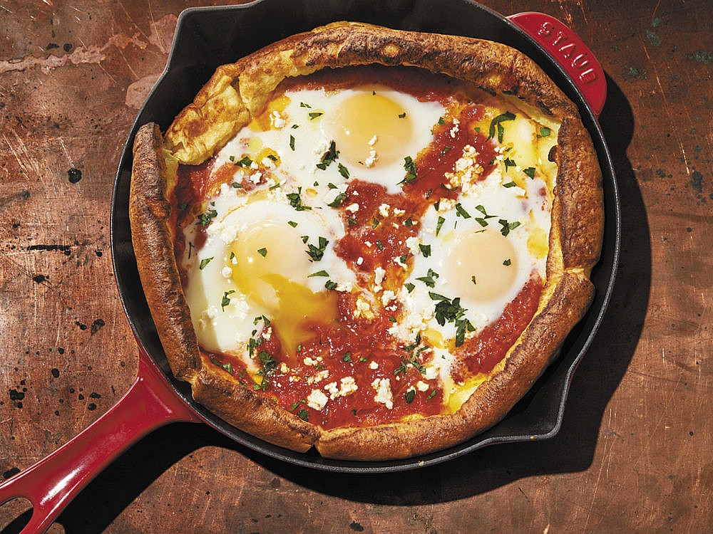 A pan with a Dutch baby filled with eggs and tomato sauce resting on a brown wooden work surface.