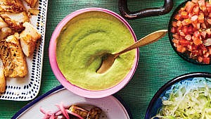 A pink bowl with green cilantro tahini sauce in it, with a gold spoon resting in the bowl, on a teal tablecloth surrounded by other taco-related dishes