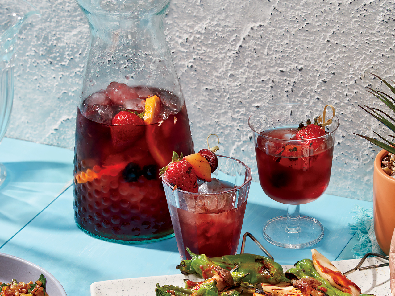 A pitcher of fruit-filled sangria with ice and a tumbler with a skewer of strawberries and fruit and sangri and a stemmed glass with the same skewr in the sangria on a blue table against a white stucco wall
