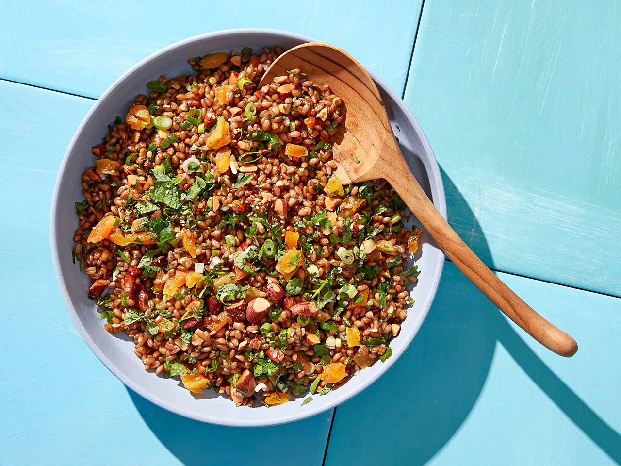 Herbed wheat berry salad with apricots and almonds in a blue bowl on a blue table with a wooden spoon resting in the salad
