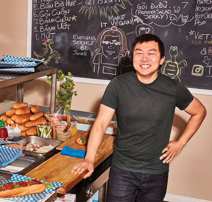 Chef Joseph Nguyễn standing by a banh mi counter in front of a blackbaoard with drawings and menu items