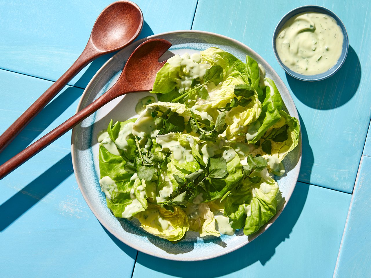Leafy greens with avocado ranch dressing on a blue and white pottery plate with wood salad tongs at the side and a bowl of buttermilk dressing on a blue table