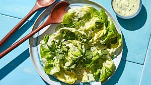 Leafy greens with avocado ranch dressing on a blue and white pottery plate with wood salad tongs at the side and a bowl of buttermilk dressing on a blue table