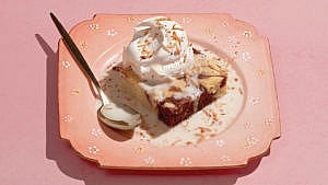 marble cake with whipped cream on top and liquid around the base of the cake, placed in a pink square plate with a metal spoon on the side of the plate.