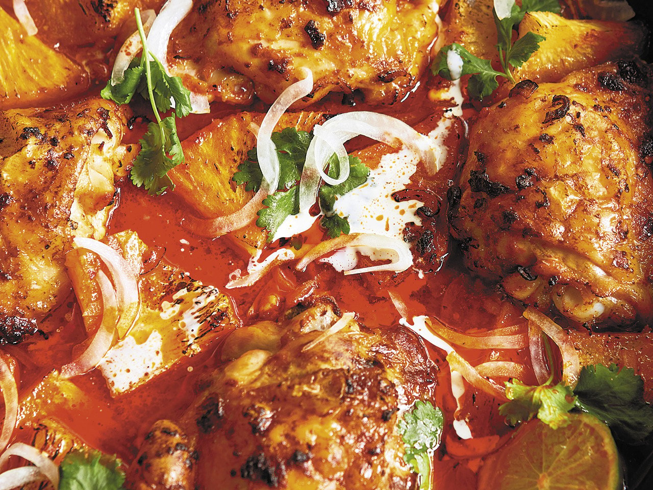 close up of cooked chicken pieces tossed in brown red sauce with drizzles of white sauce and green leaves on top.
