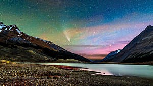 a picture of a snow-capped mountain with a lake in front at night with a comet and stars visible in the night sky