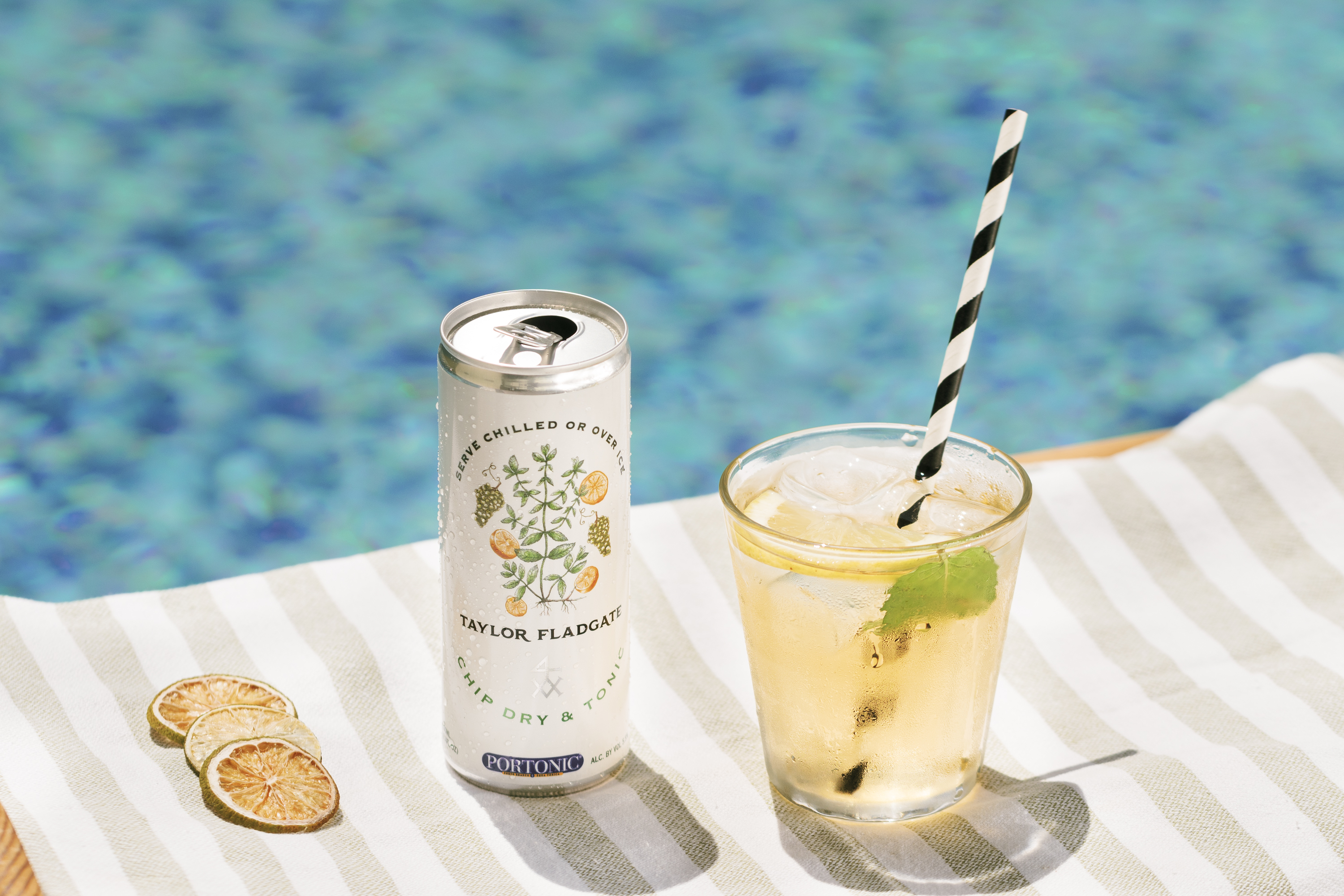 A can of Taylor Fladgate Chip Dry and Tonic in front of a pool beside a glass with the drink and a black and white striped straw and a lime slice