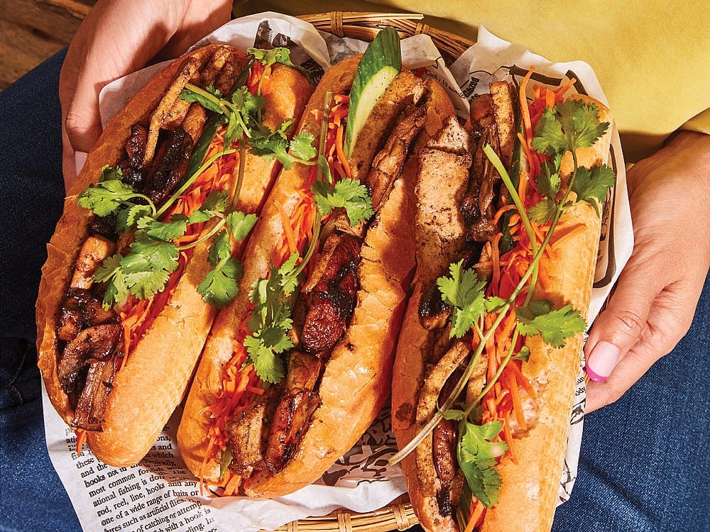 Woman holding three banh mi sandwiches that are partially open in a tray