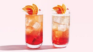 A highball glass with ice cubes and a wooden toothpick with three orange slices filled with a reddish vermouth, aquavit, Campari, sparkling wine, and orange juice cocktail on a pink background