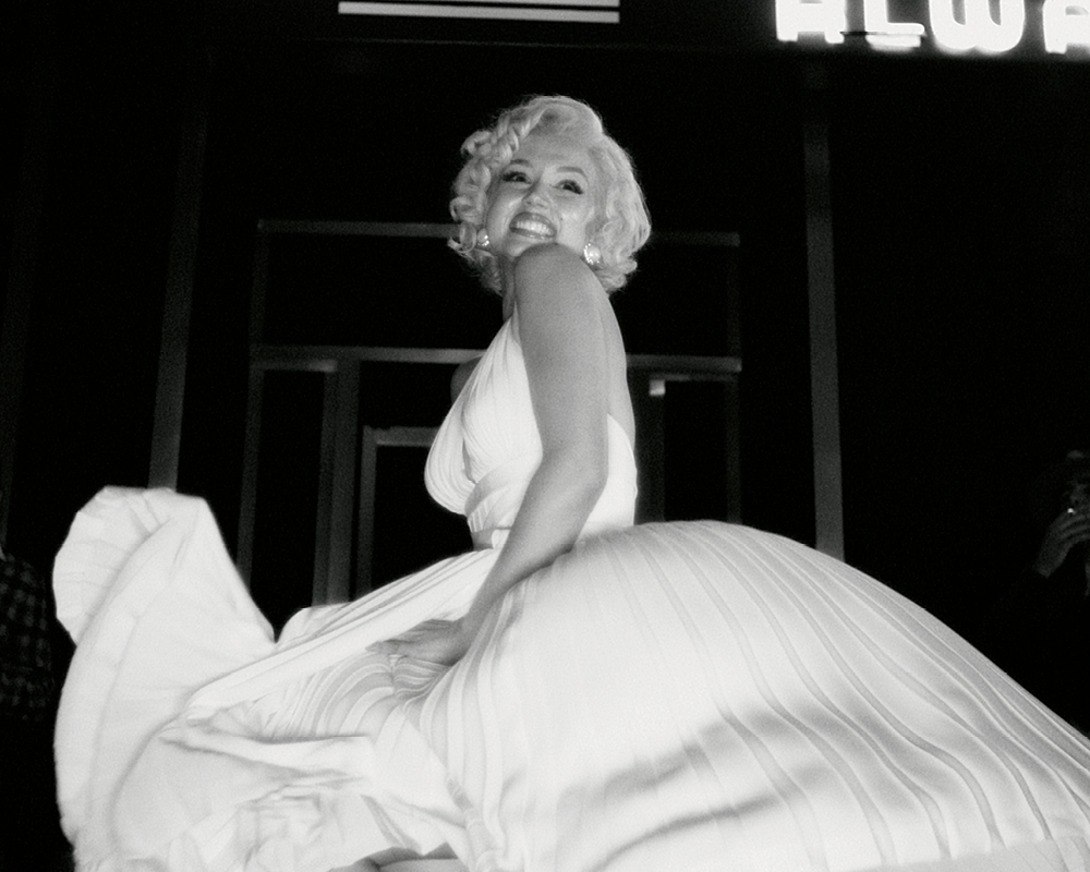 A black and white film still from Blonde, showing actress Ana de Armas as Marilyn Monroe wearing a white dress and smiling