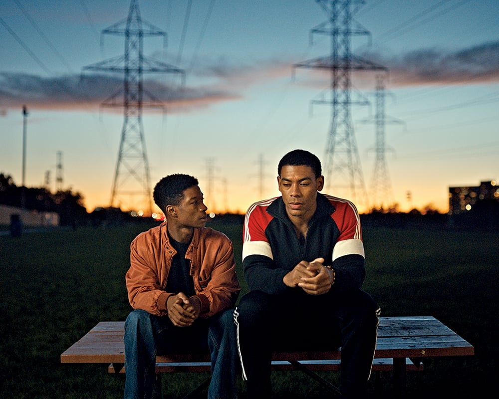 A film still from the movie Brothers, of two men sitting and speaking in fading light with phone towers in the background
