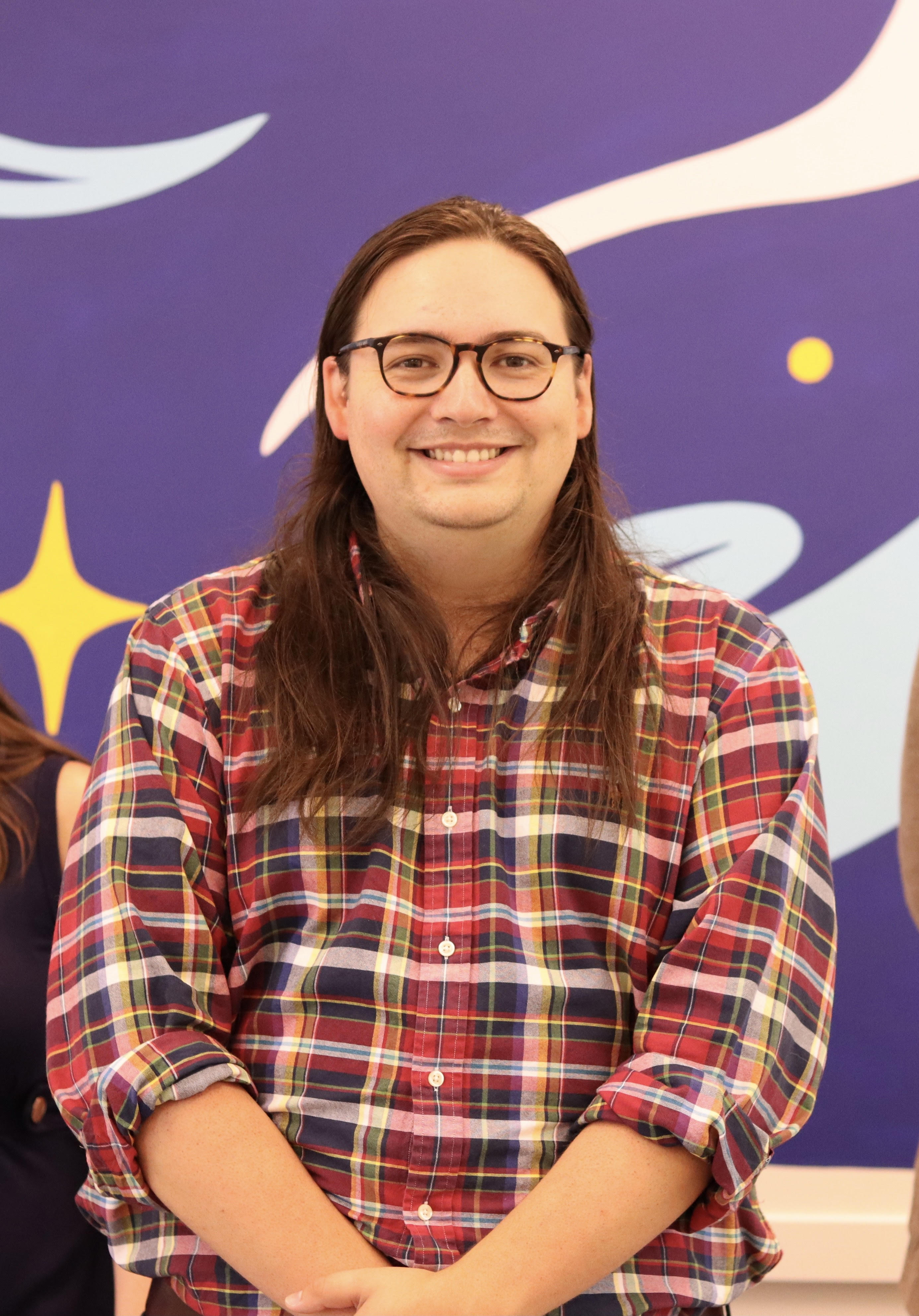 A photograph of a person with long hair and glasses, wearing a red plaid shirt and smiling into the camera