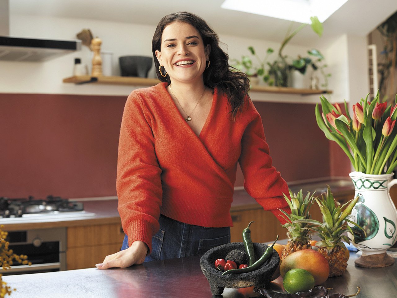 A portrait of cookbook author Ixta Belfrage standing in front of a counter with a mortar and pestle, pineapples and vase of tulips