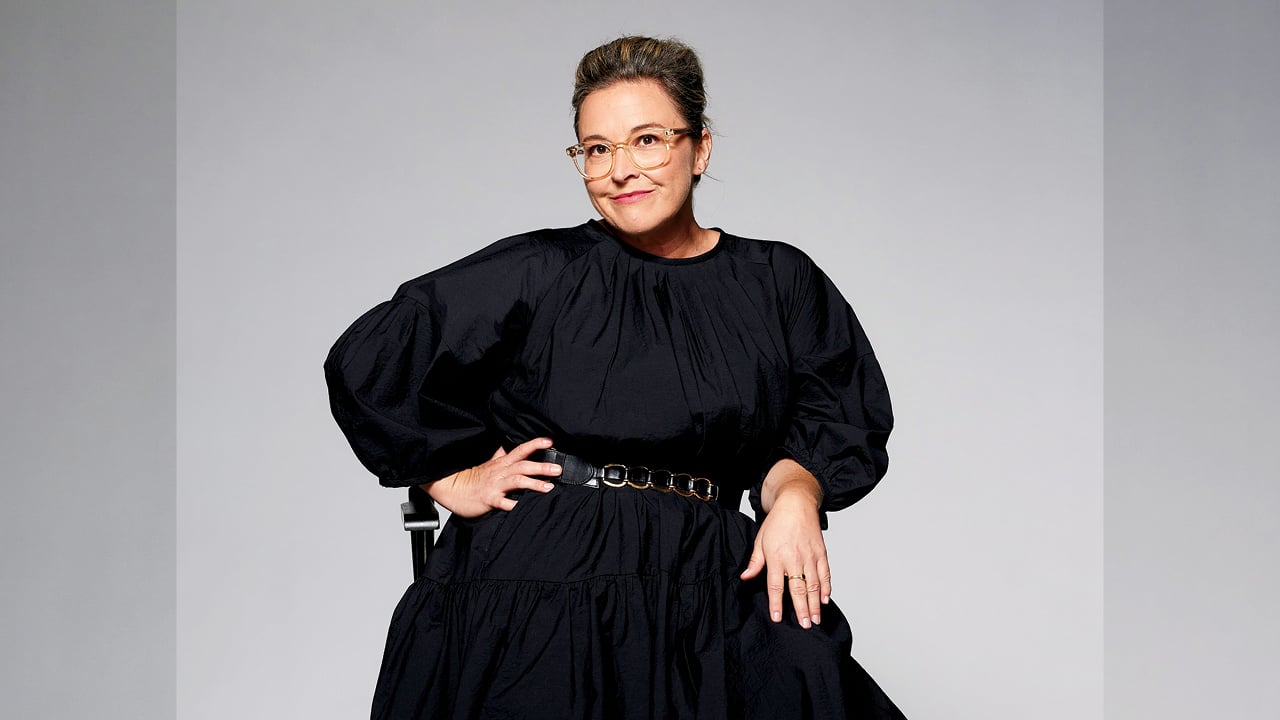 A photo of a woman with pulled back brown hair, in glasses, wearing a black dress with puffy sleeves