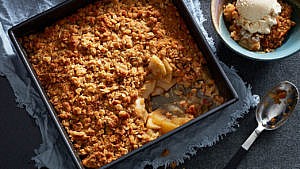 A pan of apple crisp with oats with a scoop out of it beside a bowl of apple crisp à la mode beside a spoon on a blue cloth