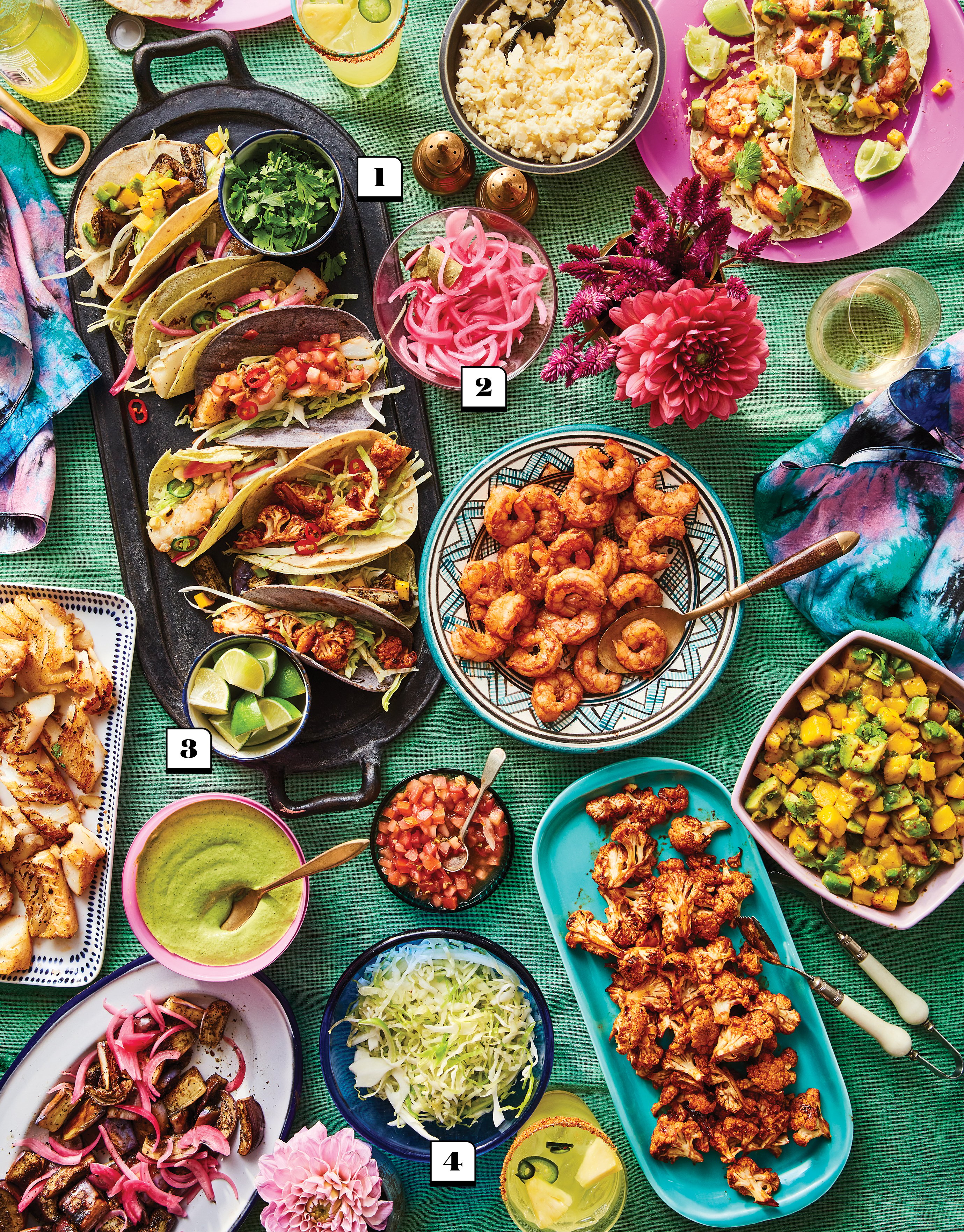 An array of tacos, drinks, and components, including pickled red onions, shrimp, salsa and shredded cabbage, laid out on a teal tablecloth