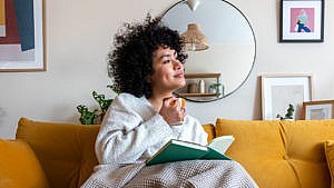 A woman sits on a couch with a coffee reading a green book
