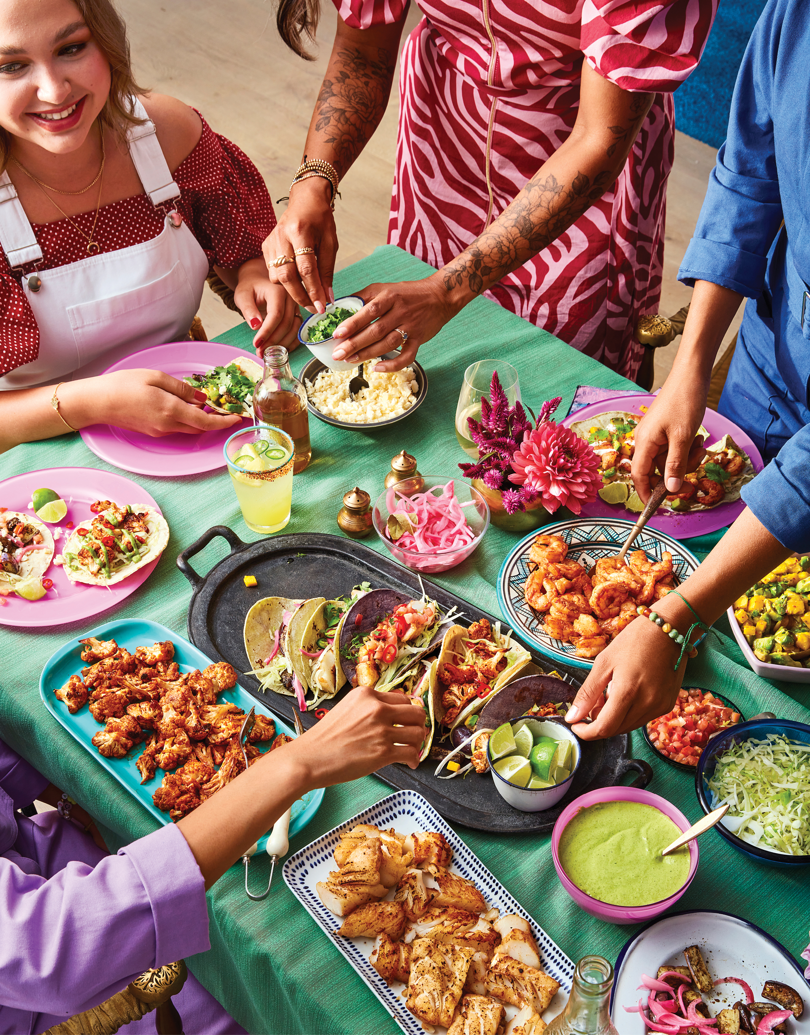 A cropped image of a table with a teal tablecloth laid out with a variety of taco dishes and fillings, and cropped to show parts of the diners sitting or standing around the table, with hands reaching in to serve