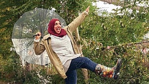 Writer Shireen Ahmed poses with a clear umbrella on a rainy day, smiling and dancing in the rain.