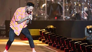 Adam Levine wearing a pink short-sleeved button down, hunched over his microphone while performing on stage