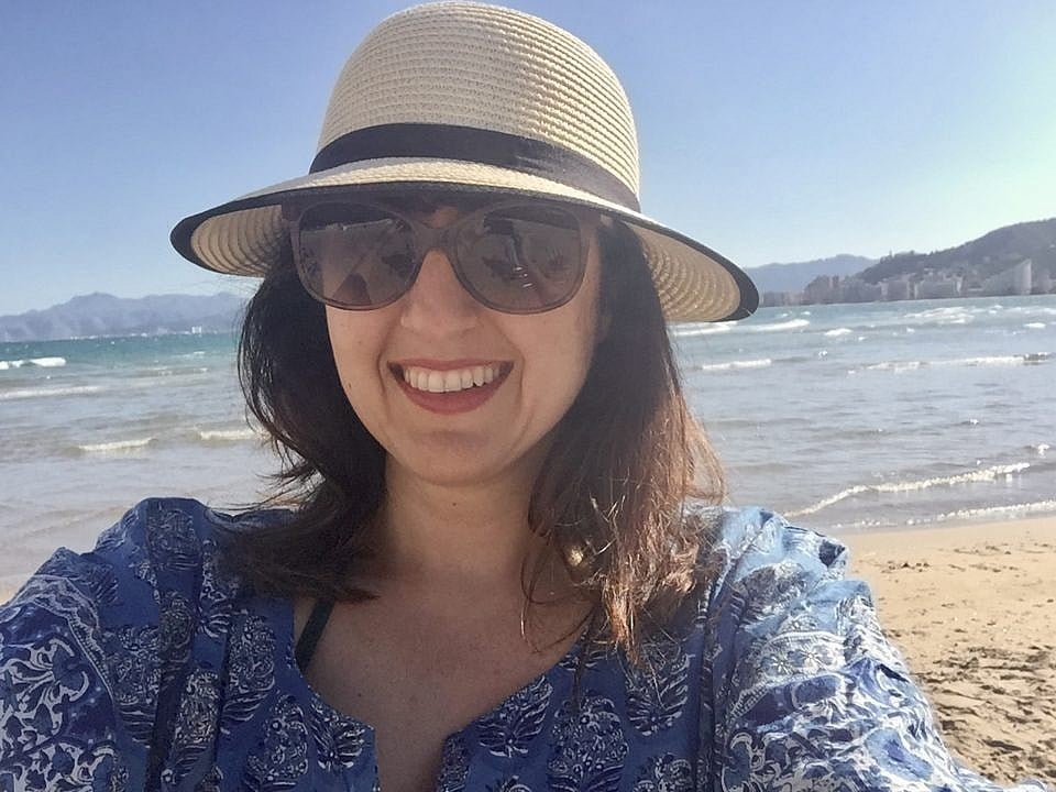 A selfie of a woman at the beach, wearing sunglasses and smiling.
