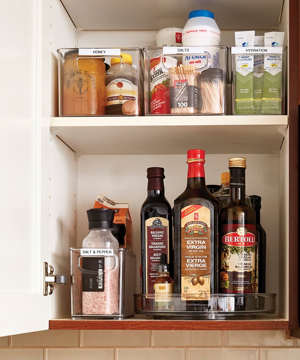 Sarah Nicole Landry's organized kitchen cupboard, with doors open to show clear bins storing condiments and spices, and a clear lazy susan turntable holding bottles of oils and vinegars.