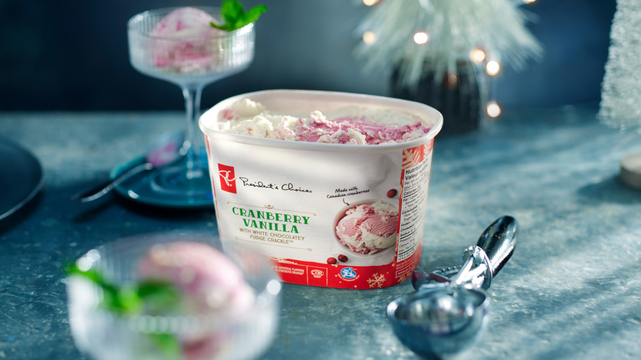 A tub of PC Cranberry Vanilla with White Chocolatey Fudge Crackle Ice Cream next to bowls and scoop