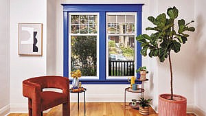 A window with vivid blue trim, surrounded by a red velvet chair, end tables and a plant.