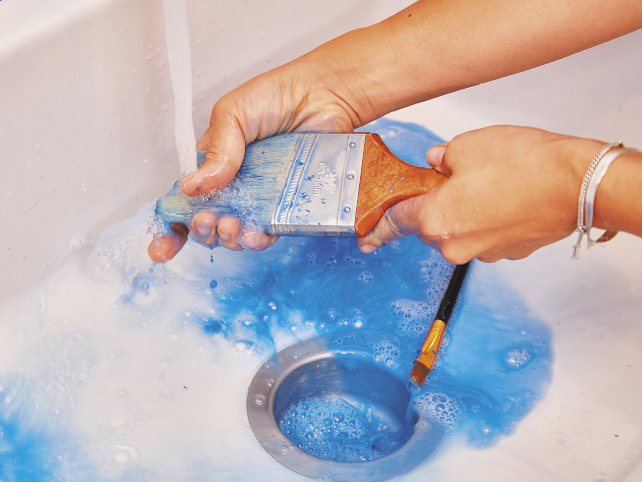 A pair of hands cleaning blue paint off a paint brush over a sink with running water.