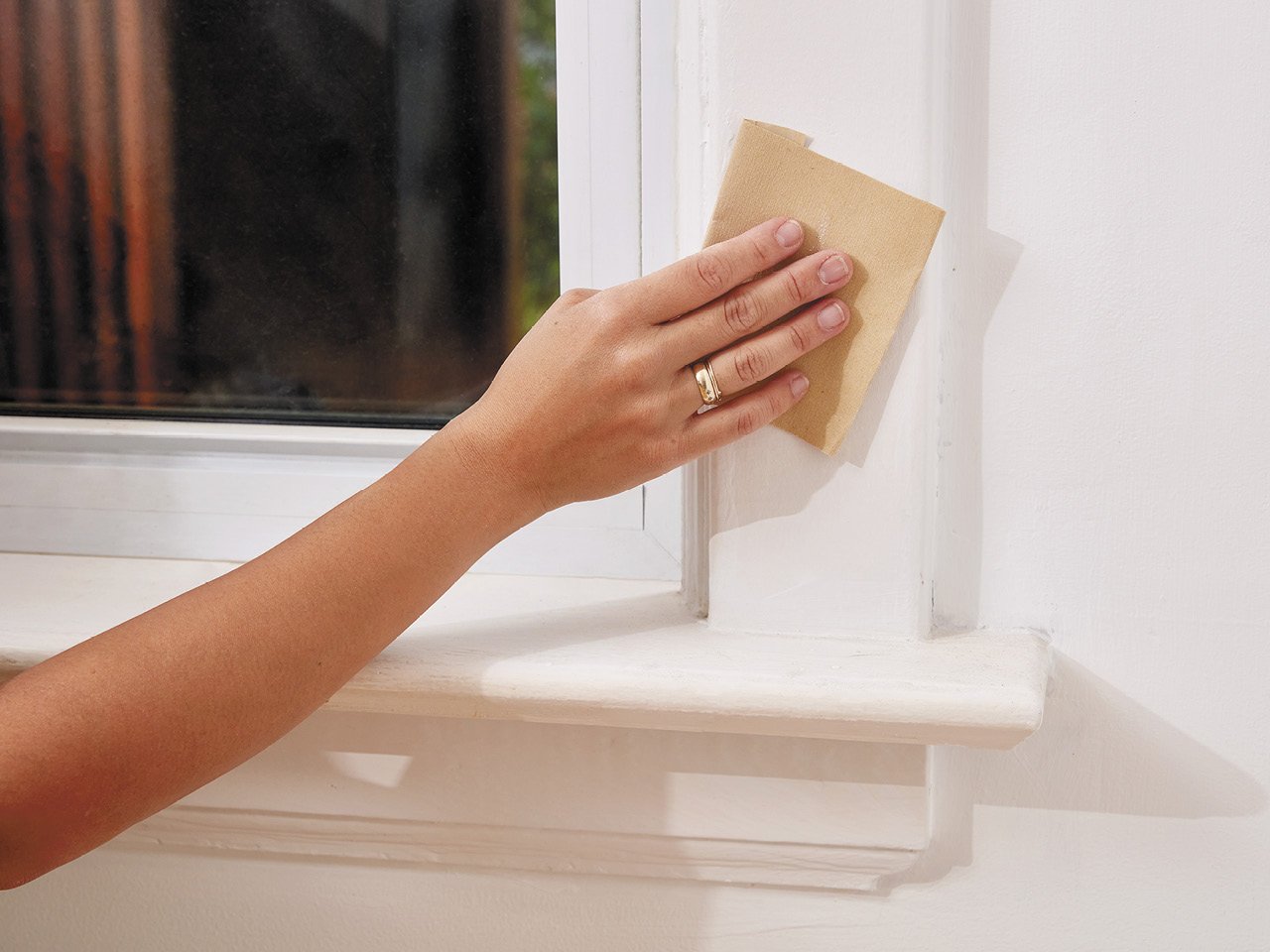 A hand sanding a white window trim to prep it for a coat of blue paint.