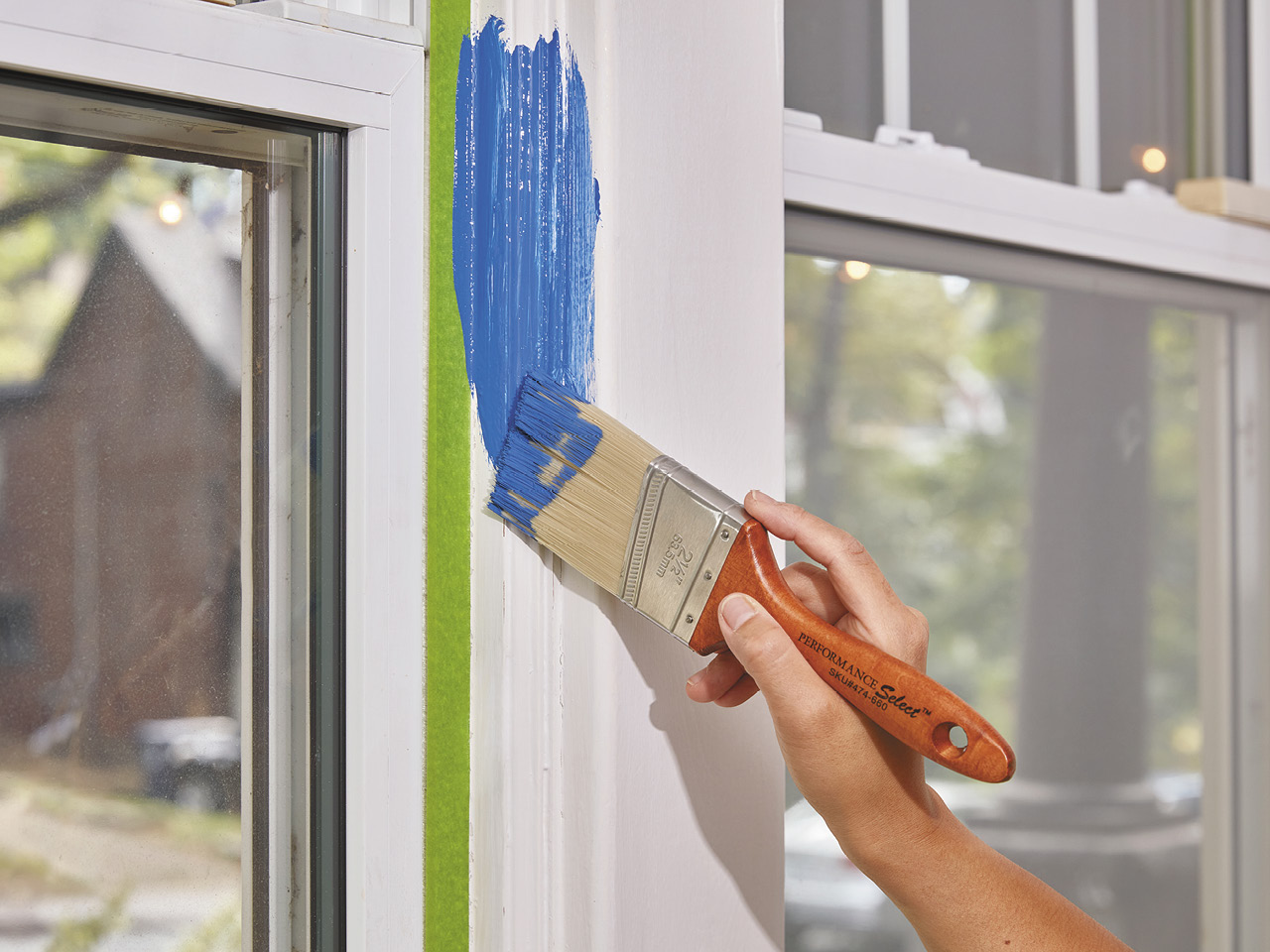 A hand holding a paint brush applying blue paint on a white window trim.