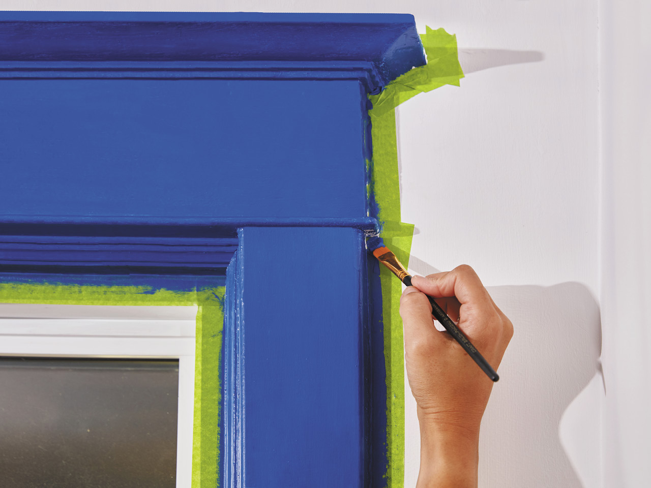 A hand holding a small paint brush touching up a painted blue window trim. 