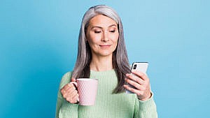A woman with grey hair stands against a blue backdrop, holding a phone in her left hand and a mug in her right hand