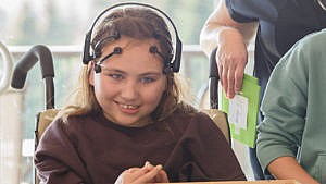 A young girl in a wheelchair wearing a computerized headset, using Brain Computer Interface (BCI) technology as a communication pathway for children with severe speech and motor impairments.