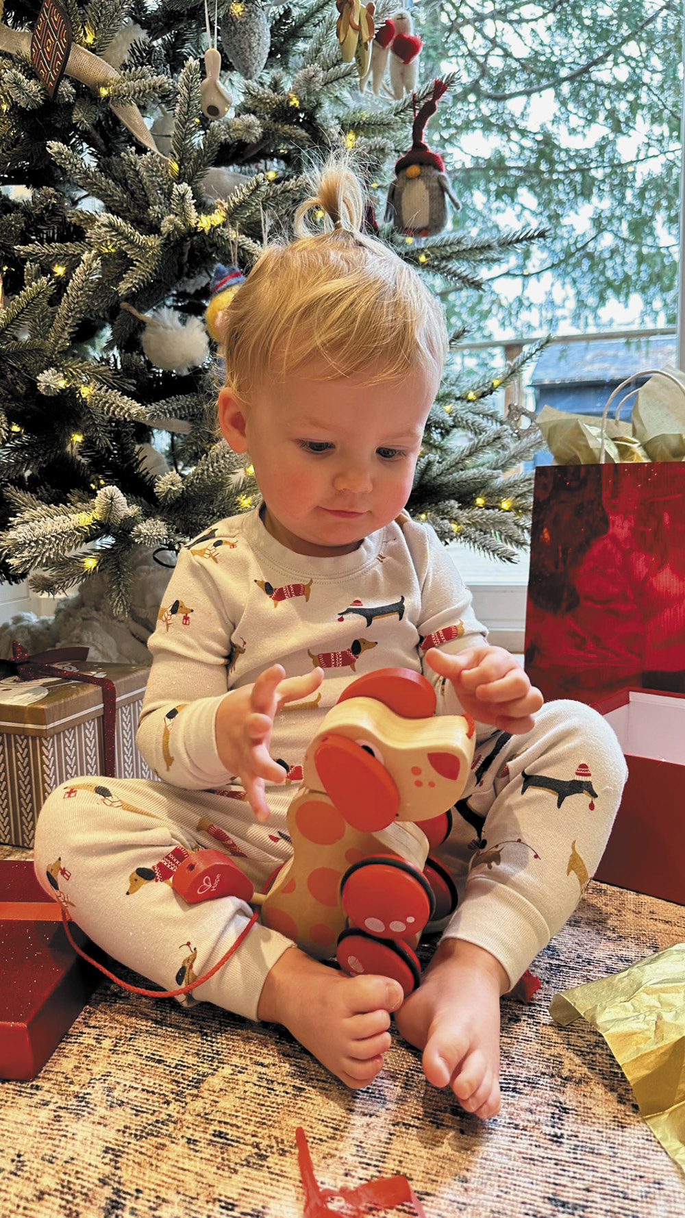 A toddler playing with a wooden dog underneath a Christmas tree.