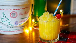 a glass of yellow pineapple slush with a cherry and paper straw