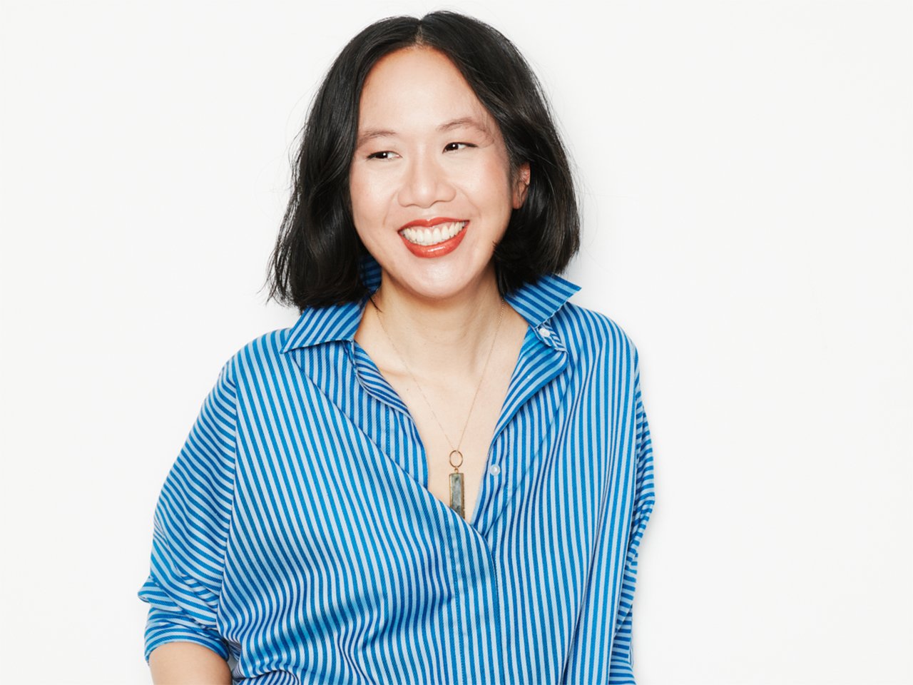 Author Jen Sookfong Lee on a white background, smiling and wearing a blue vertical striped blouse