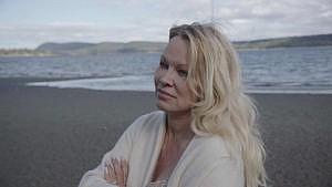 Pamela Anderson stands on a beach with the ocean behind her, wearing a white sweater and crossing her arms